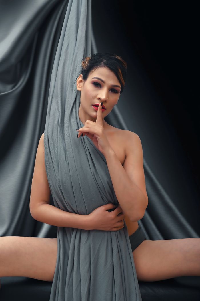 A woman draped in fabric making a silence gesture, exuding elegance indoors.